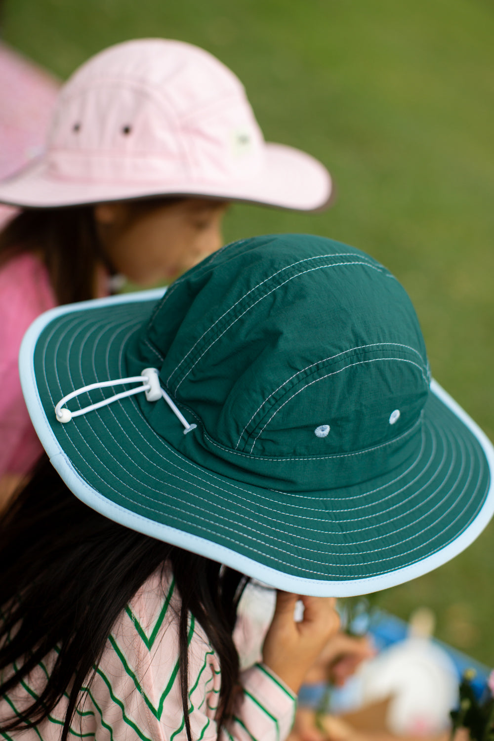 A close-up of a child wearing a Beeji Earth Child dark green wide-brim sun hat with an adjustable drawstring and blue trim, with another child in a pink sun hat blurred in the background on a grassy field.