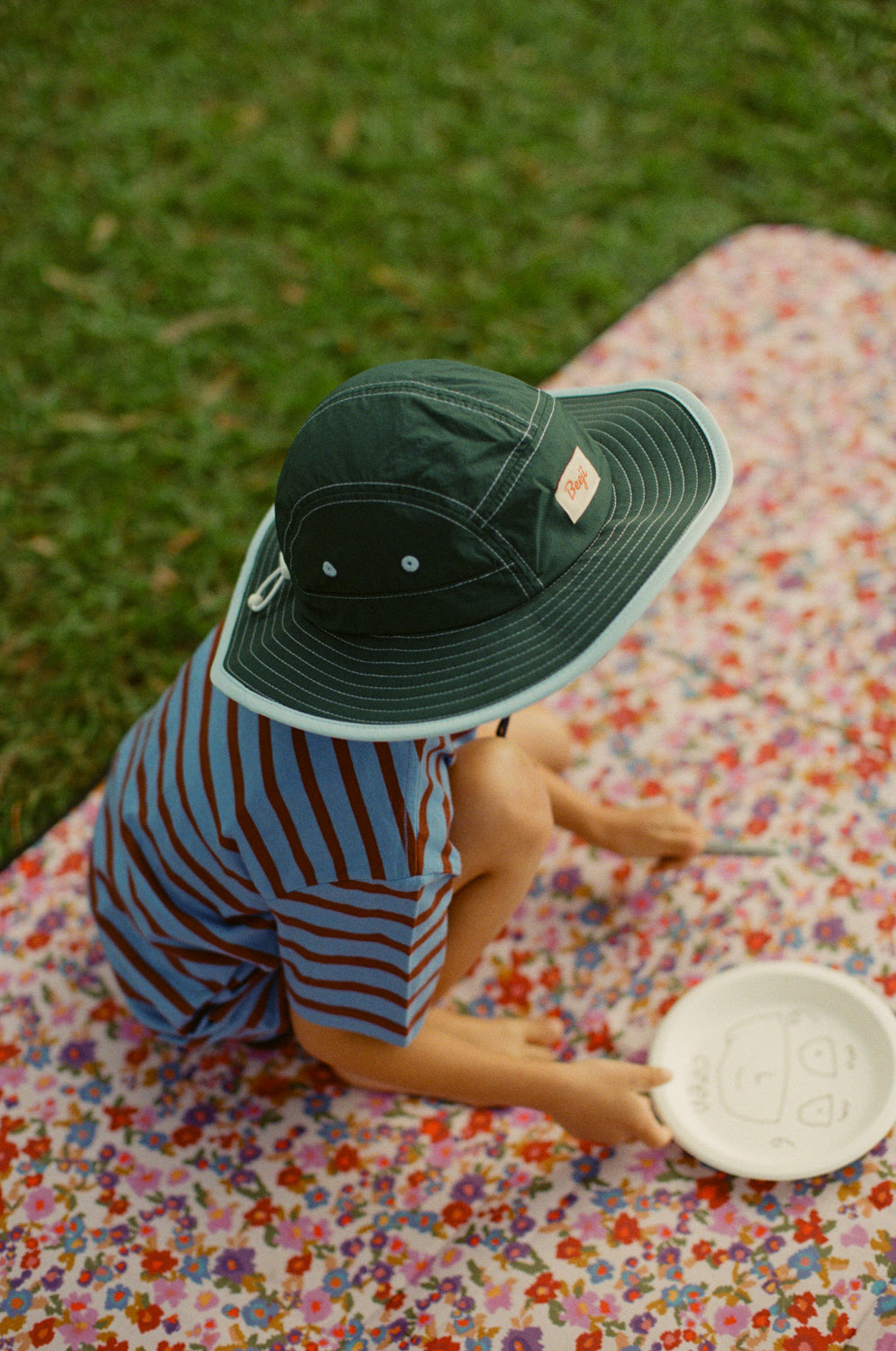 A child sitting on a floral picnic blanket outdoors on grass, wearing a Beeji Earth Child dark green wide-brim sun hat and blue trim and a blue-and-red striped shirt, holding a white plate.